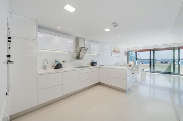 A modern kitchen with white cabinetry, a built-in oven, and a view of the sea through large windows. - Ático Londres Derecha a Modern 2º floor Penthouse Beachfront in Puerto Pollensa (Puerto Pollensa)