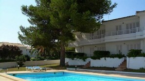 Una piscina con tumbonas, un árbol grande y un edificio blanco con balcones.
