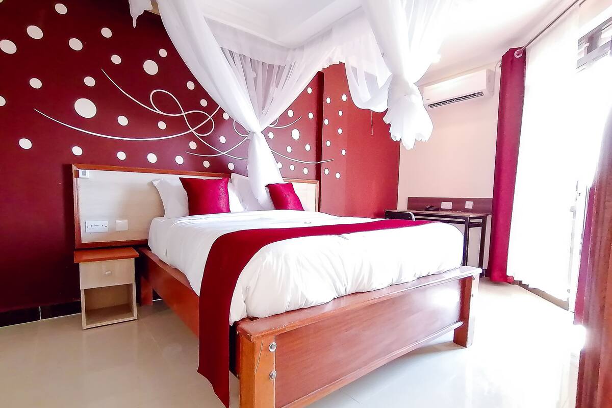 Quarto com cama de madeira, mesa de cabeceira, espelho e parede vermelha com bolinhas brancas.