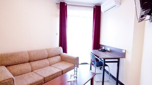 A small, well-lit room with a sofa, a wooden table, and a desk with a chair. There is a window with curtains, an air conditioning unit on the wall, and a wall-mounted TV. - Divine Homes Resort (Kisumu)