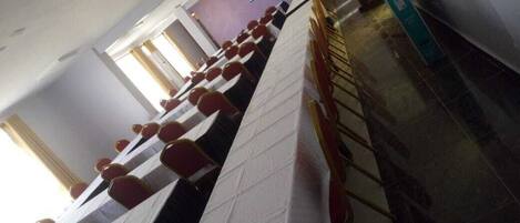 A conference room with rows of tables and chairs, a television screen on the wall, and a window with curtains.