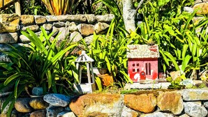 Un jardín con muro de piedra, diversas plantas y una pequeña casa roja en miniatura.