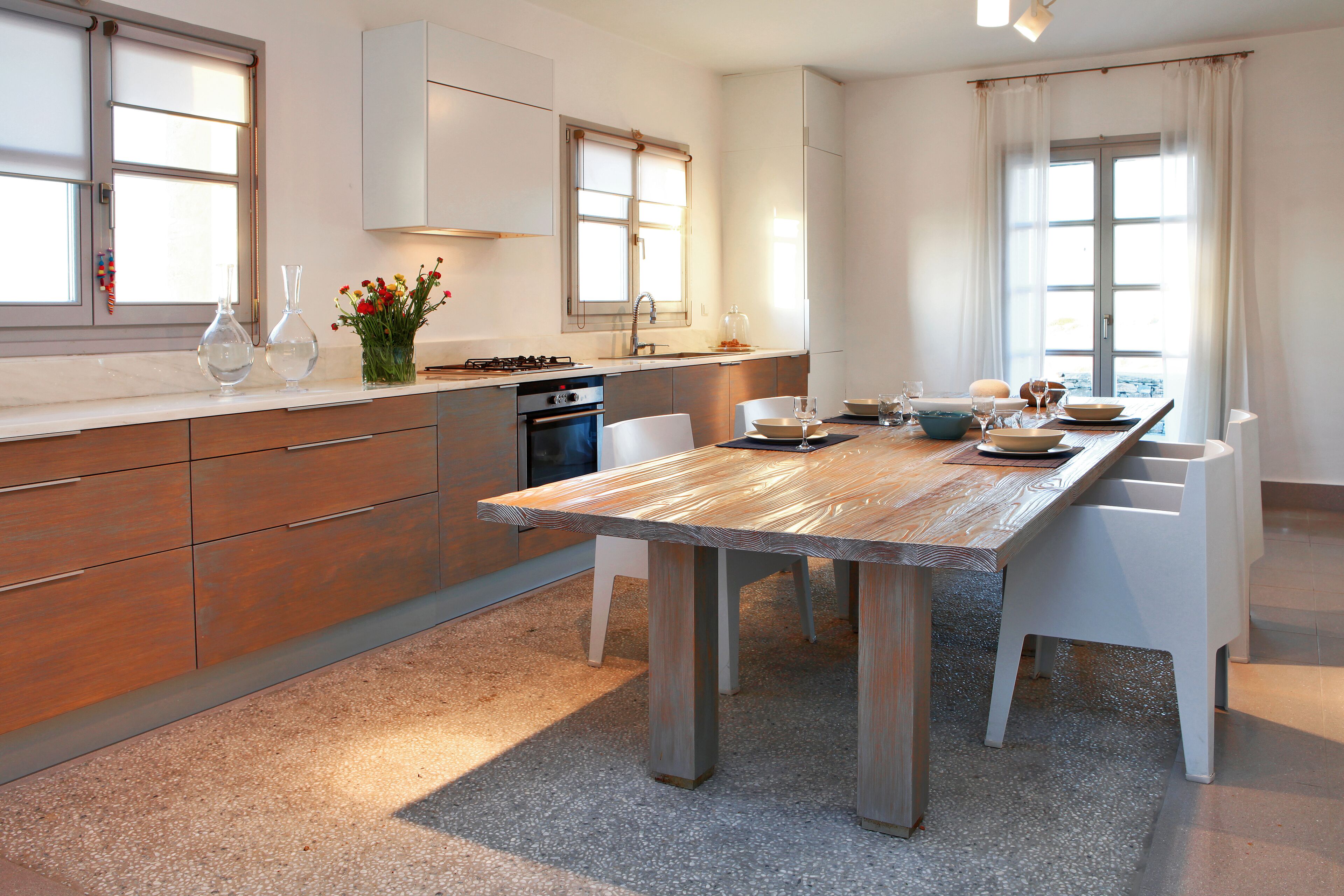 A modern kitchen with a wooden table, white chairs, and a large window.