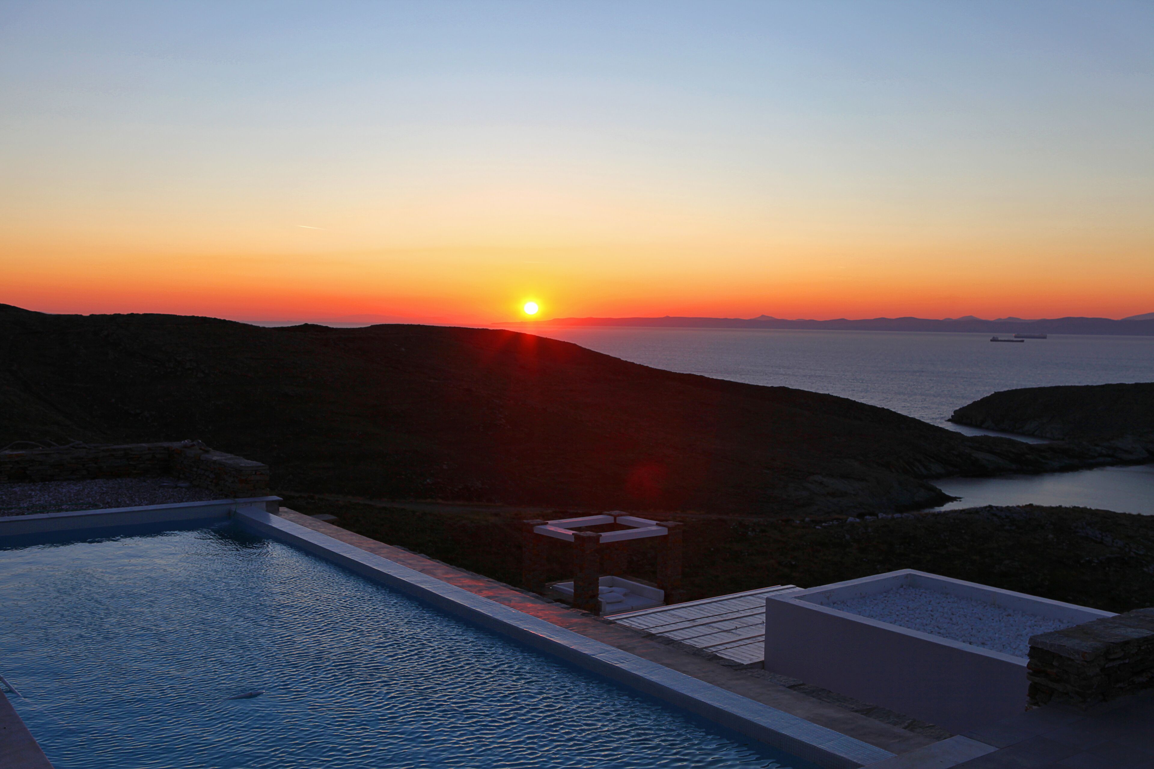 Une vue sur le coucher de soleil depuis un espace aménagé près de la piscine, avec vue sur la mer et les collines lointaines.
