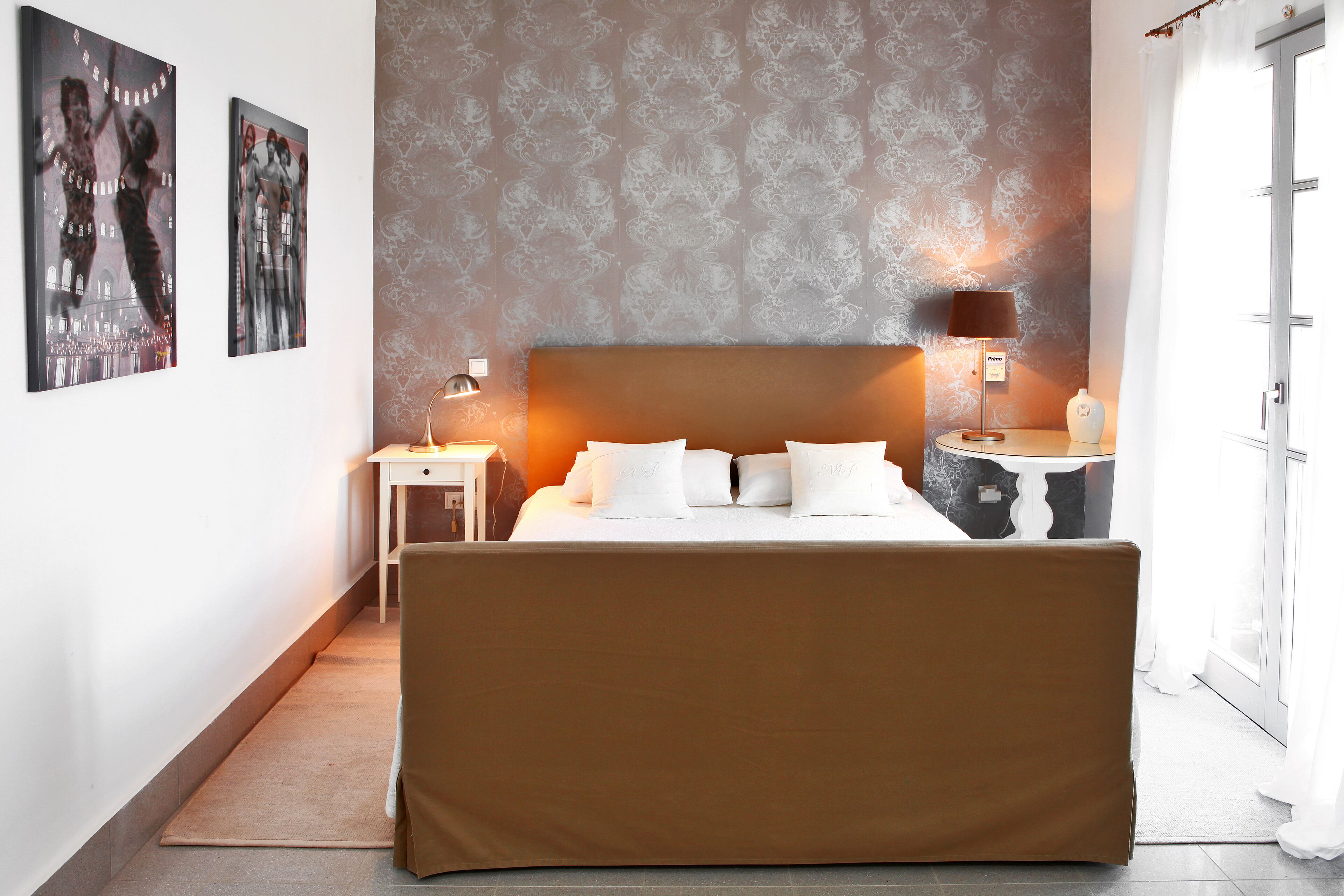 A hotel room with a large bed, a nightstand with a lamp, a wall with patterned wallpaper, and two framed pictures on the wall.
