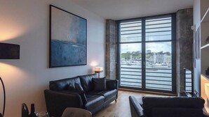 Un salon moderne avec un canapé en cuir noir, une table à manger avec des chaises, une grande fenêtre donnant sur un port et un tableau accroché au mur.