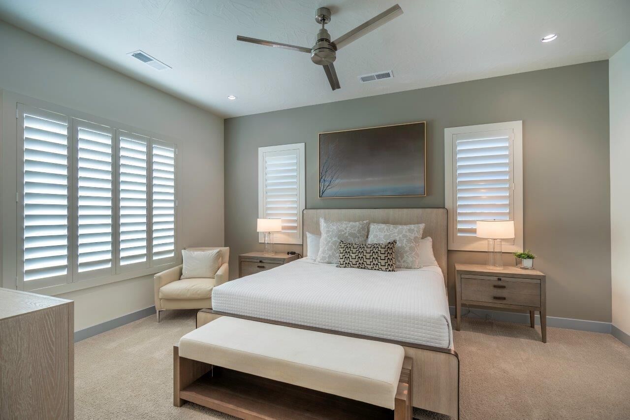 A bedroom with a bed, two bedside tables, a chair, a ceiling fan, and large windows with blinds.