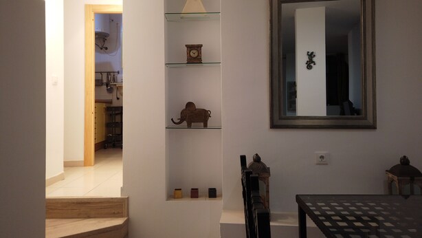 Una habitación con una escalera que lleva a una cocina, un espejo montado en la pared y estantes con objetos decorativos.