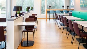 Un espace repas moderne avec des tables et des chaises en bois, de grandes fenêtres donnant sur la verdure et un coin salon vert.