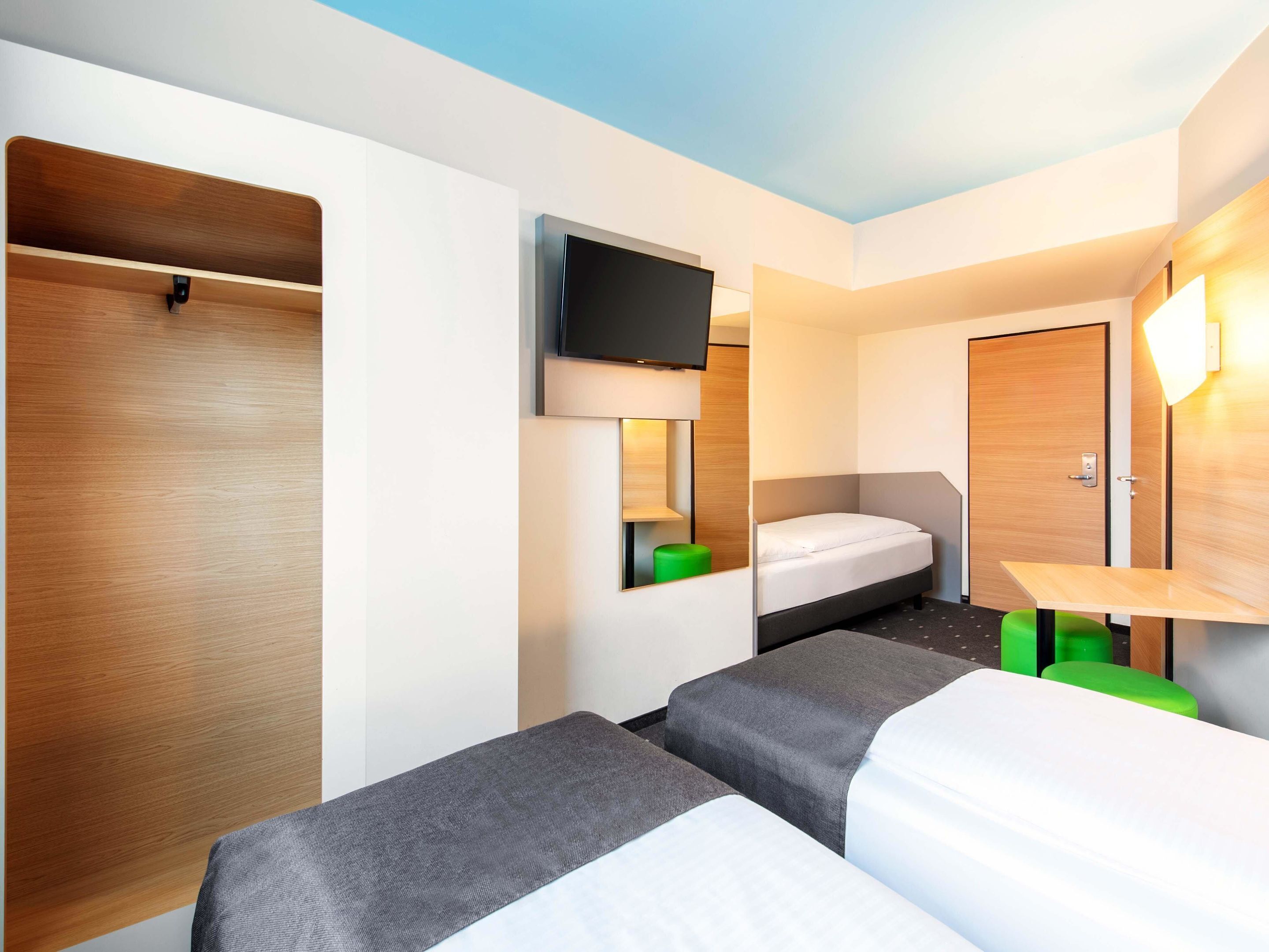 Habitación de hotel con dos camas individuales, un televisor montado en la pared, un armario de madera y una mesita con un taburete verde.