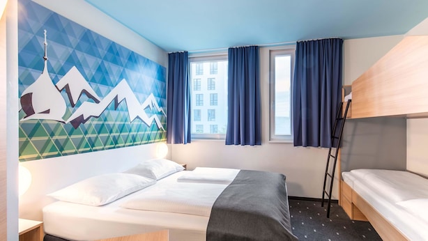 Une chambre d’hôtel comprenant un lit, un bureau, une fenêtre avec des rideaux, un mur orné d’une fresque géométrique et un lit superposé.