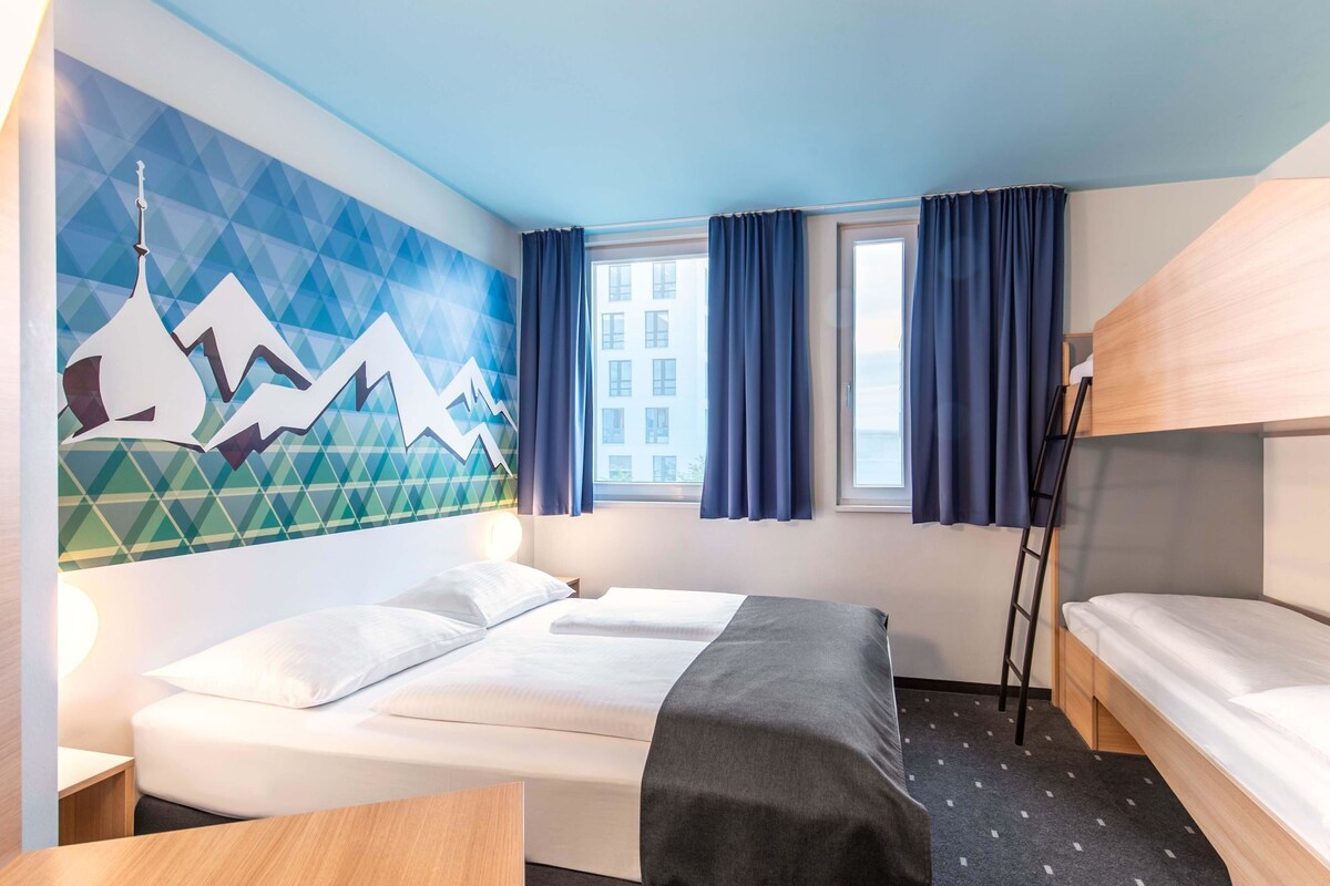 A hotel room with a bed, a desk, a window with curtains, a wall with a geometric mural, and a bunk bed.