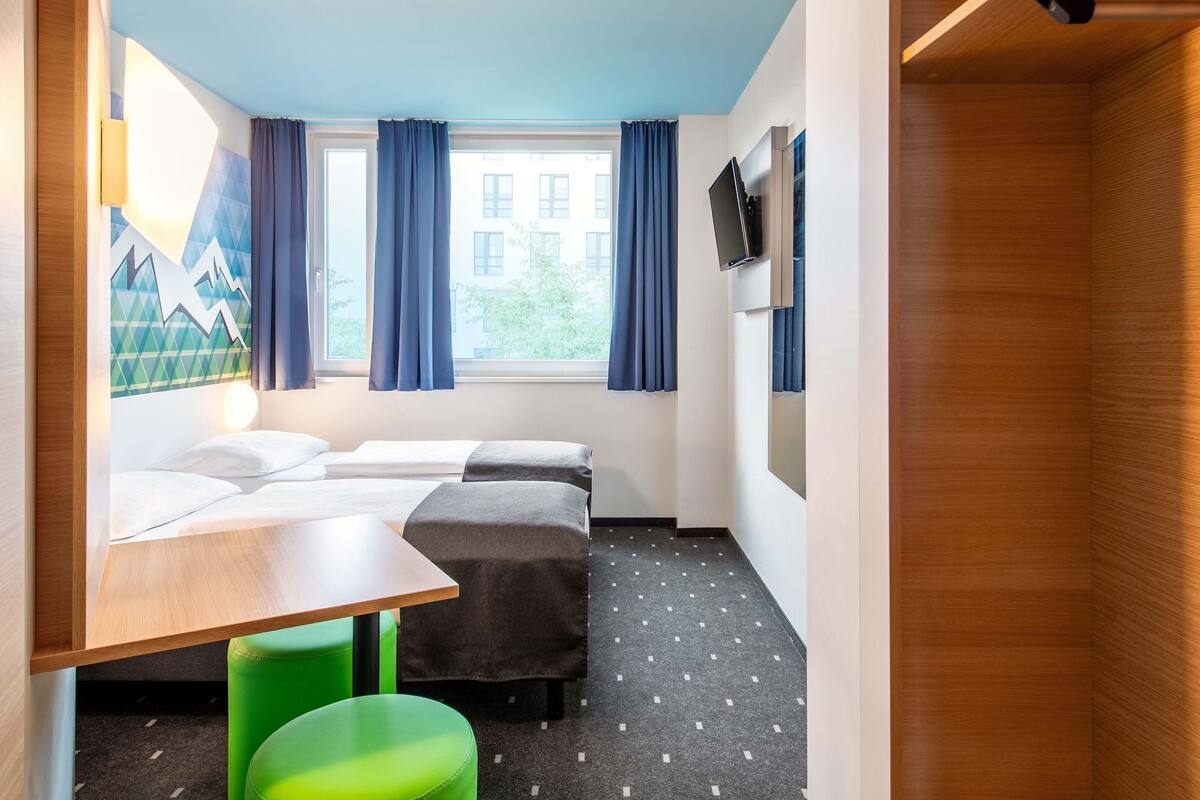 Quarto de hotel com cama, escrivaninha, um banquinho verde, televisão e janela com cortinas.
