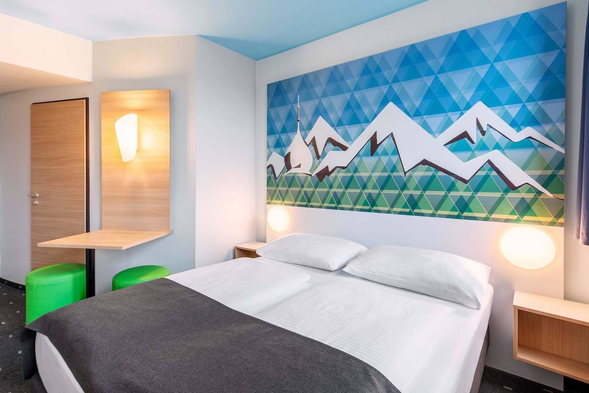 Quarto de hotel com uma cama, um banquinho verde, uma mesa de cabeceira de madeira e uma parede com um desenho geométrico de montanhas.