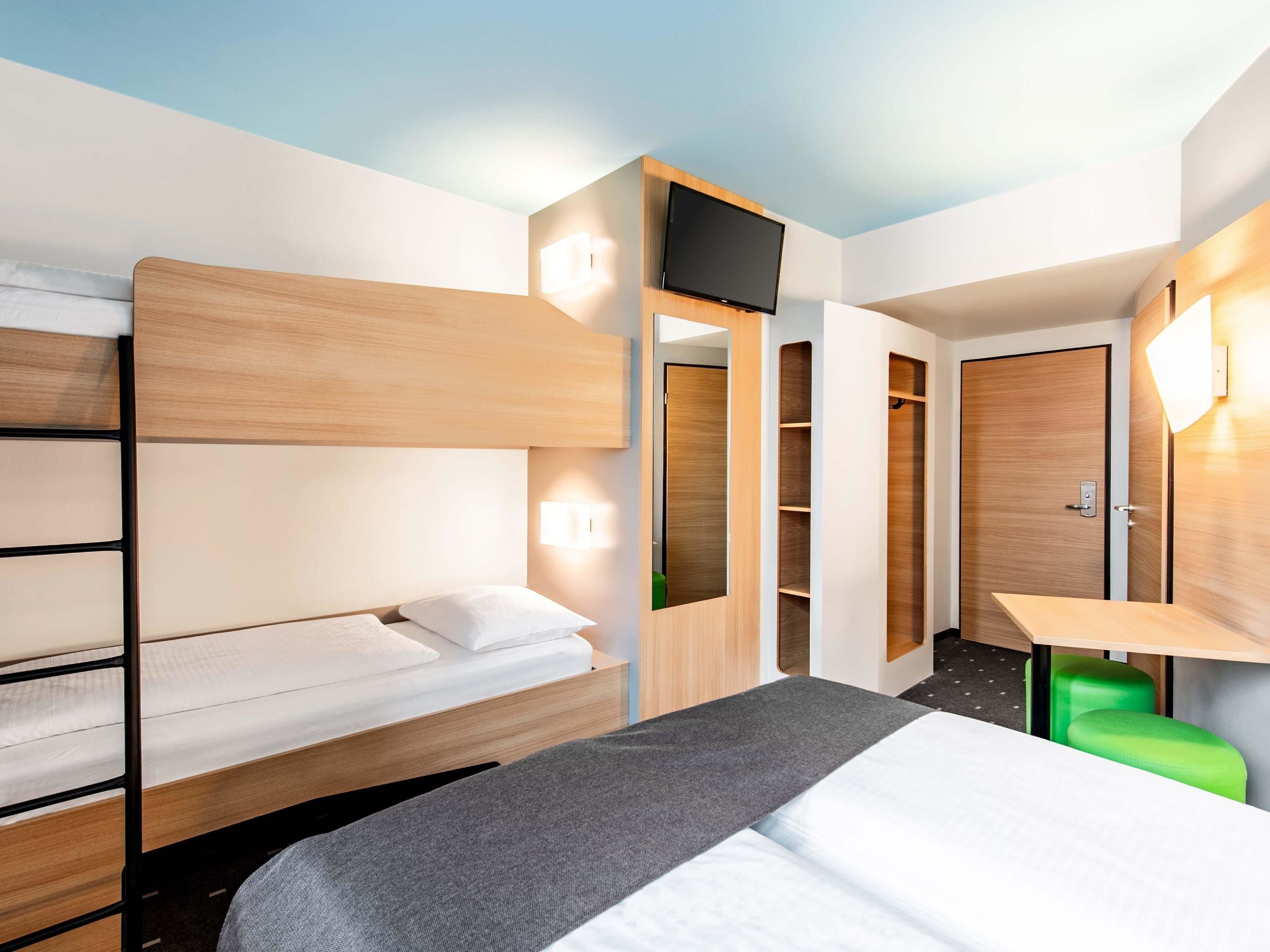 A hotel room with a bunk bed, a desk with a chair, a TV mounted on the wall, and a wardrobe with shelves.