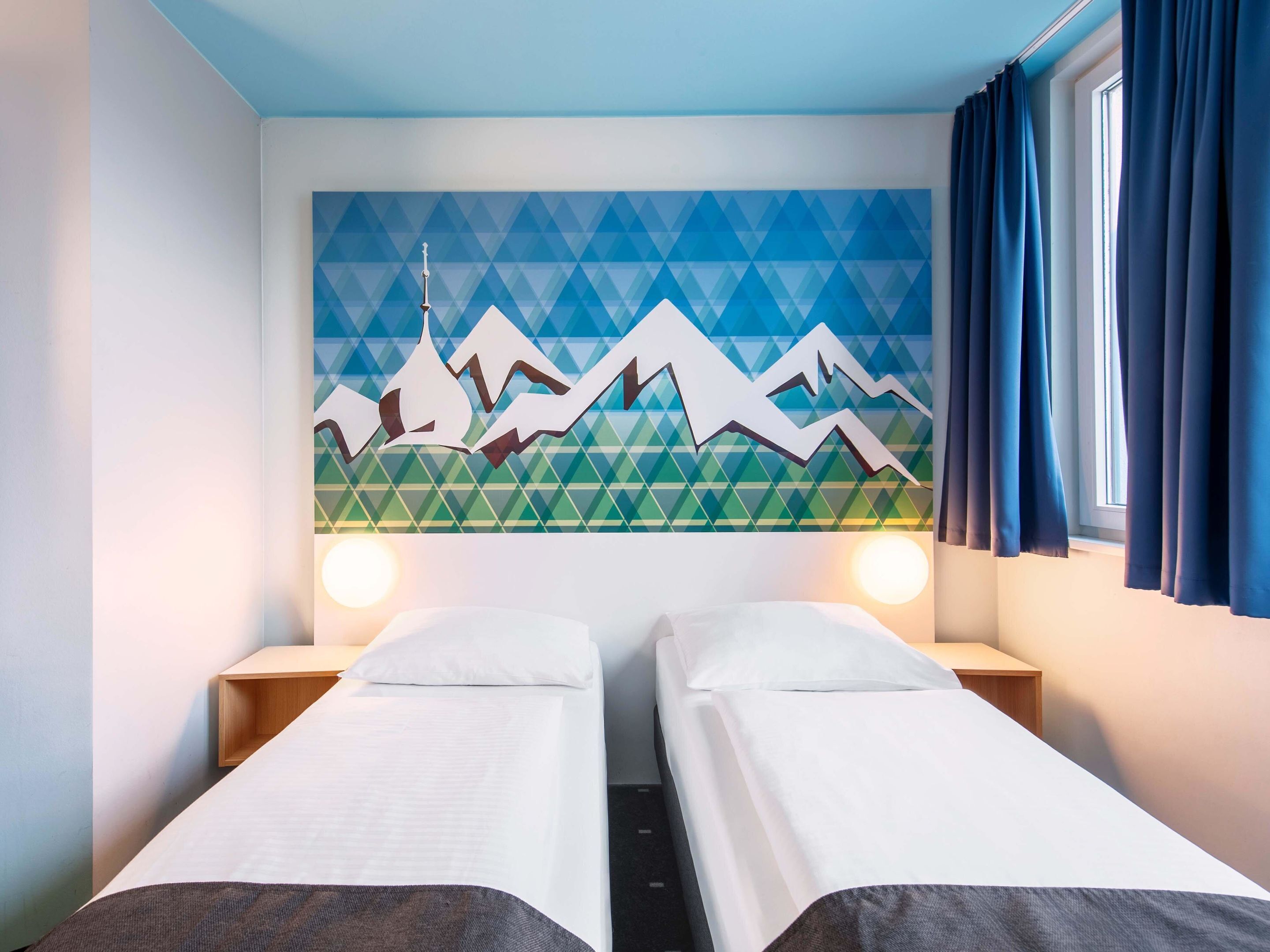 Ein Hotelzimmer mit zwei Einzelbetten, einem geometrischen Wandbild und einem Fenster mit Vorhängen.