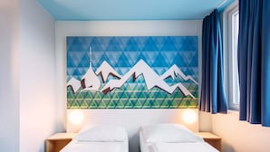 Une chambre d’hôtel avec deux lits simples, une fresque murale aux motifs géométriques et une fenêtre avec des rideaux.