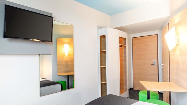 Una habitación de hotel moderna con televisión de pantalla plana, un espejo grande, un armario, un escritorio y otomanas verdes.