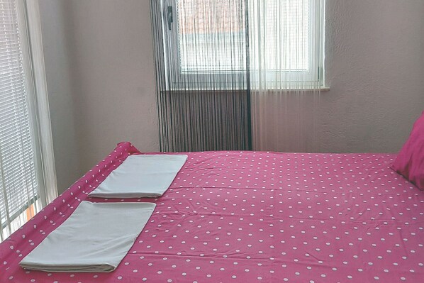 Une petite pièce avec un lit recouvert de draps à pois roses, une fenêtre avec des stores verticaux et un mur uni.