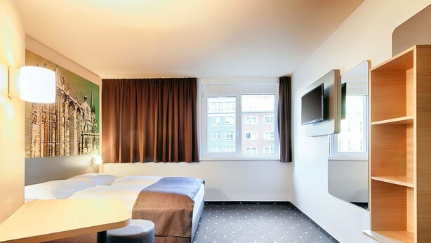 Habitación de hotel con cama, escritorio, silla, televisor y ventana con cortinas.