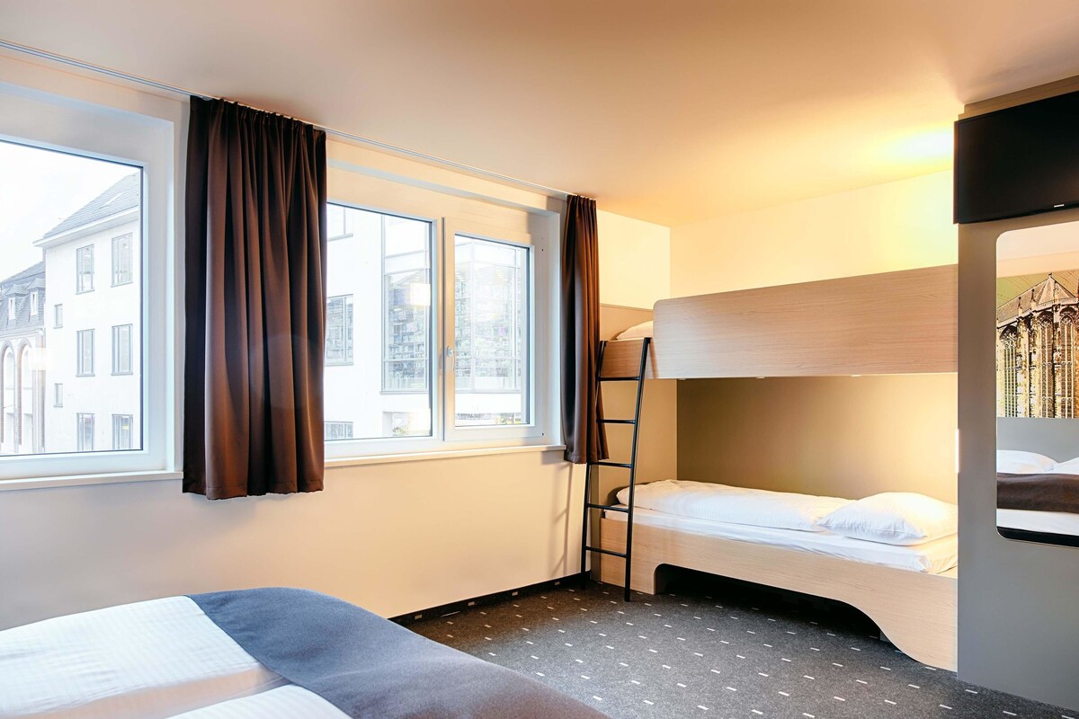 Een hotelkamer met twee bedden, een stapelbed, een raam met gordijnen, een ladder en een televisie.