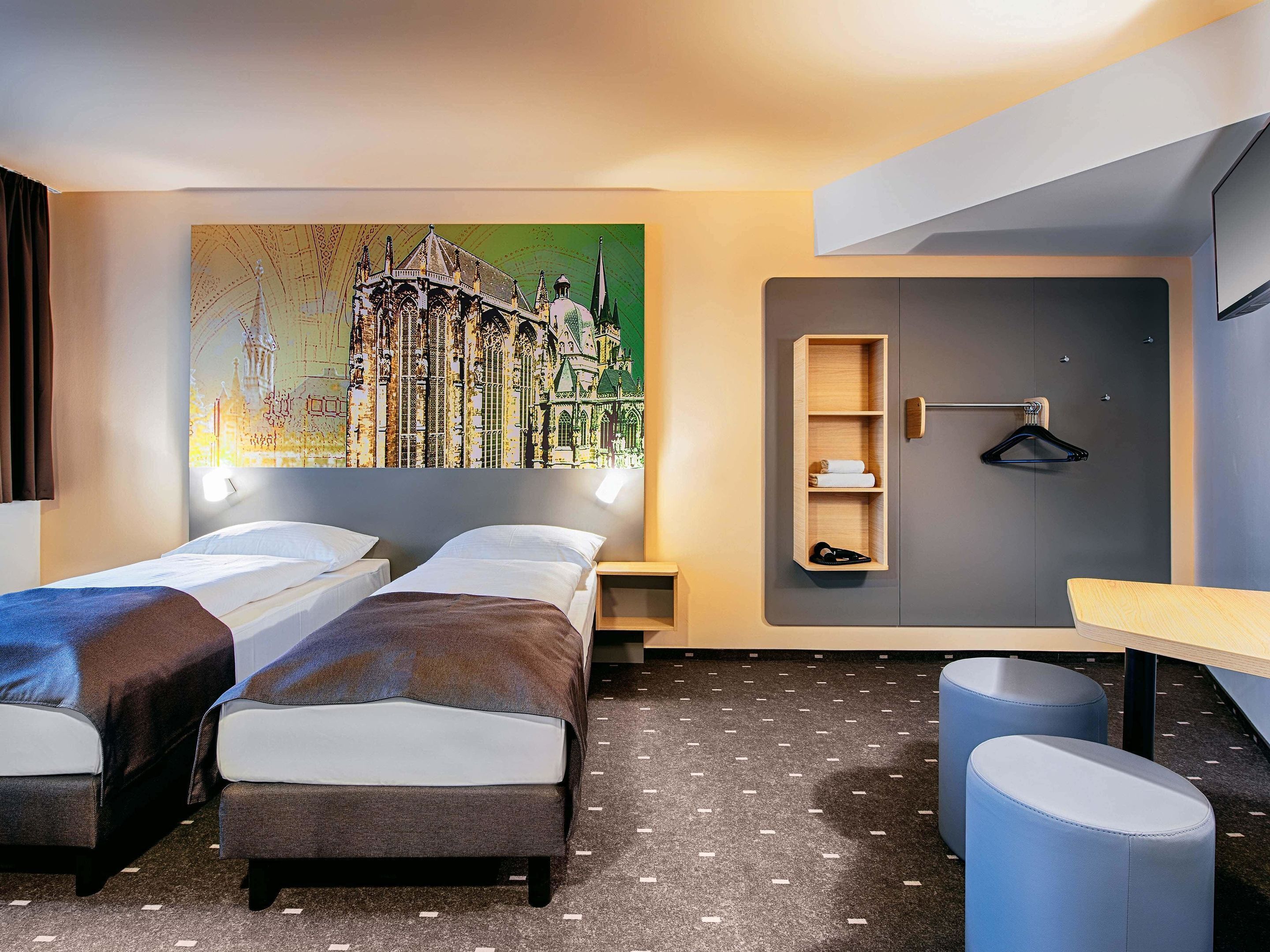 Ein Hotelzimmer mit zwei Einzelbetten, einem an der Wand befestigten Kleiderschrank, einem kleinen Tisch und einem großen Stadtbild.