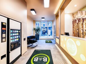 Een hotel receptie met een automaat, een receptiebalie en uitzicht op de straat door grote ramen.