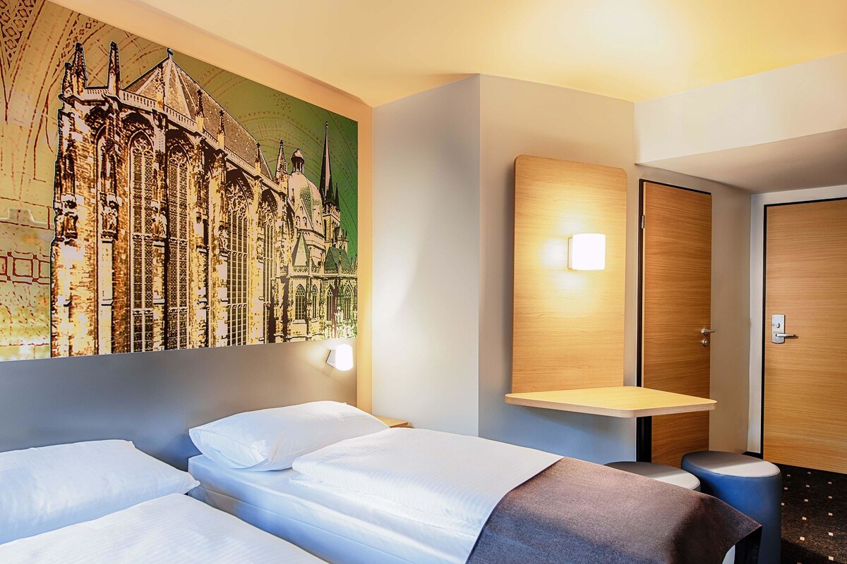 Een hotelkamer met twee bedden, een wandplank, een deur en een kunstwerk van een gebouw.
