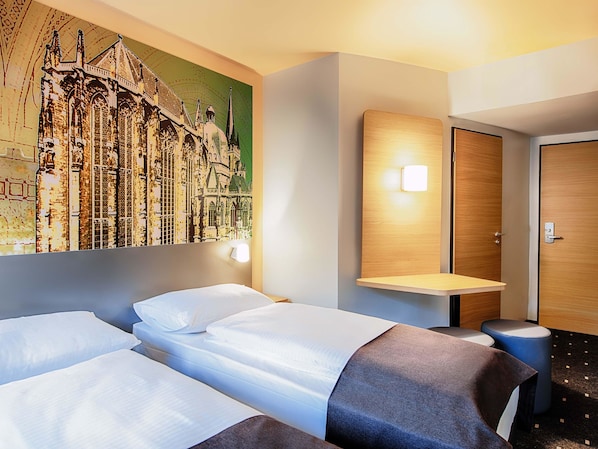 A hotel room with two beds, a wall-mounted shelf, a door, and a wall art depicting a building.