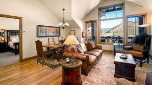 A spacious loft with a high ceiling, a large window offering a view of the outdoors, a cozy seating area with a leather sofa and armchairs, and a dining table with chairs. - Beaver Creek Landing A404 3 Bedroom Condo by RedAwning (Avon)