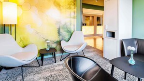 A modern hotel room with a large abstract mural, two white chairs, a black leather chair, a small table with a vase, and a reception desk in the background. - B&B Hotel Baden-Airpark (Rheinmunster)