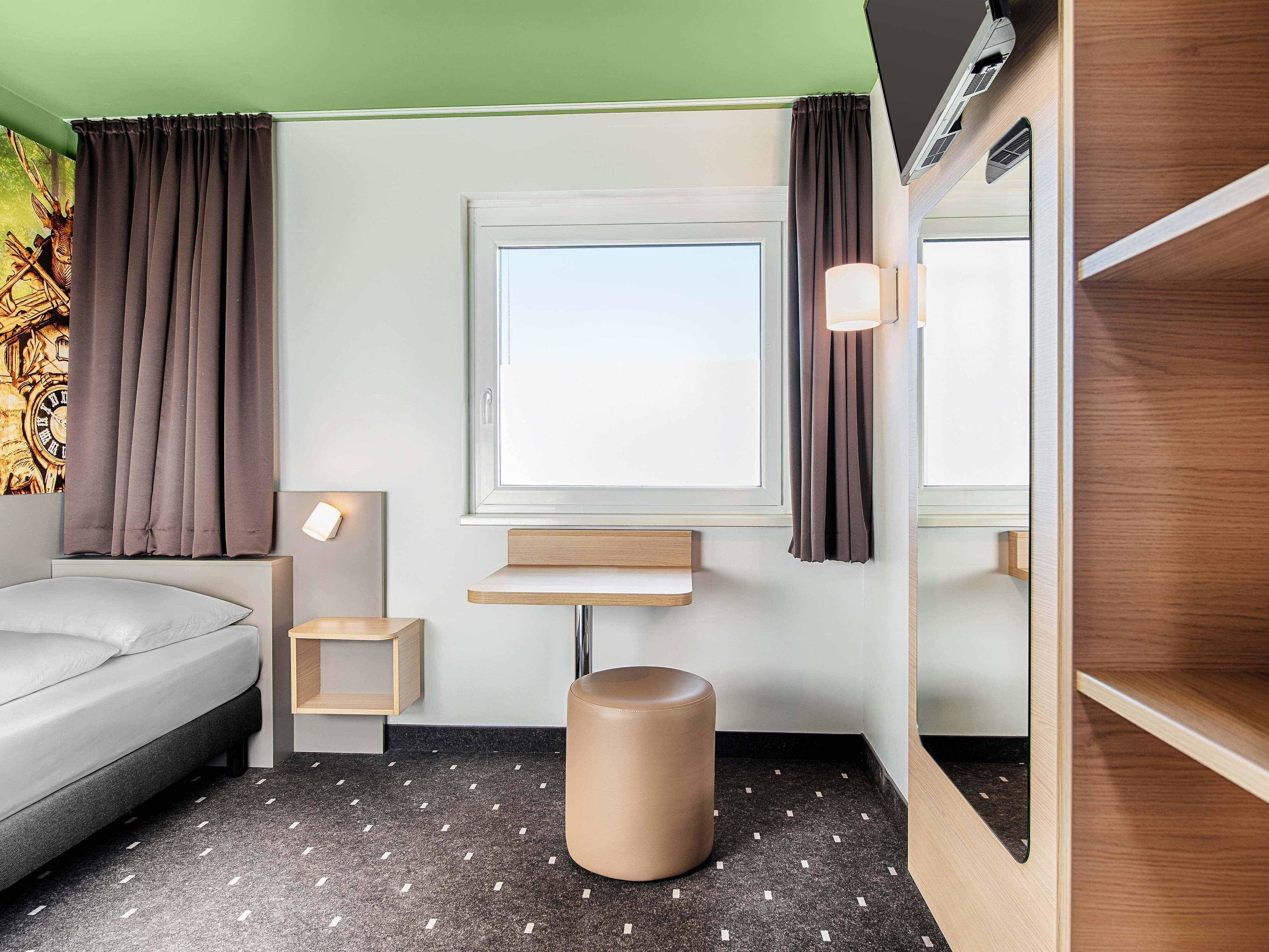 Een hotelkamer met een bed, een klein nachtkastje, een wandplank, gordijnen voor het raam en een rond krukje.