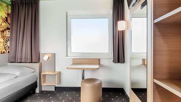 Een hotelkamer met een bed, een klein nachtkastje, een wandplank, gordijnen voor het raam en een rond krukje.