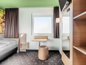 Une chambre d’hôtel comprenant un lit, une petite table de chevet, une commode, une fenêtre avec des rideaux, une étagère fixée au mur et un tabouret rond.
