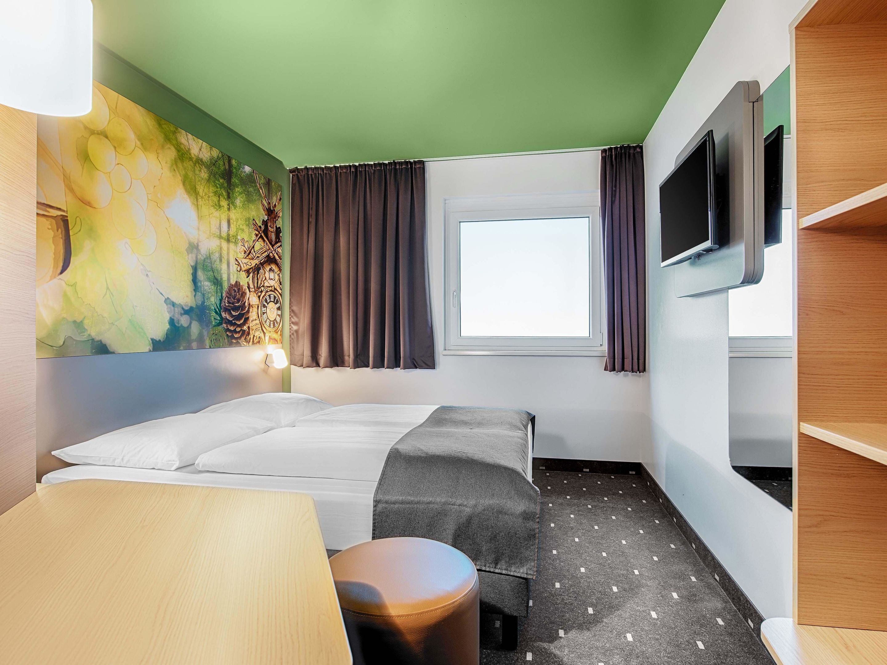 Una habitación de hotel con cama, escritorio, silla, televisor, ventana con cortinas y un mural colorido en la pared.