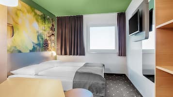 Una habitación de hotel con cama, escritorio, silla, televisor, ventana con cortinas y un mural colorido en la pared.