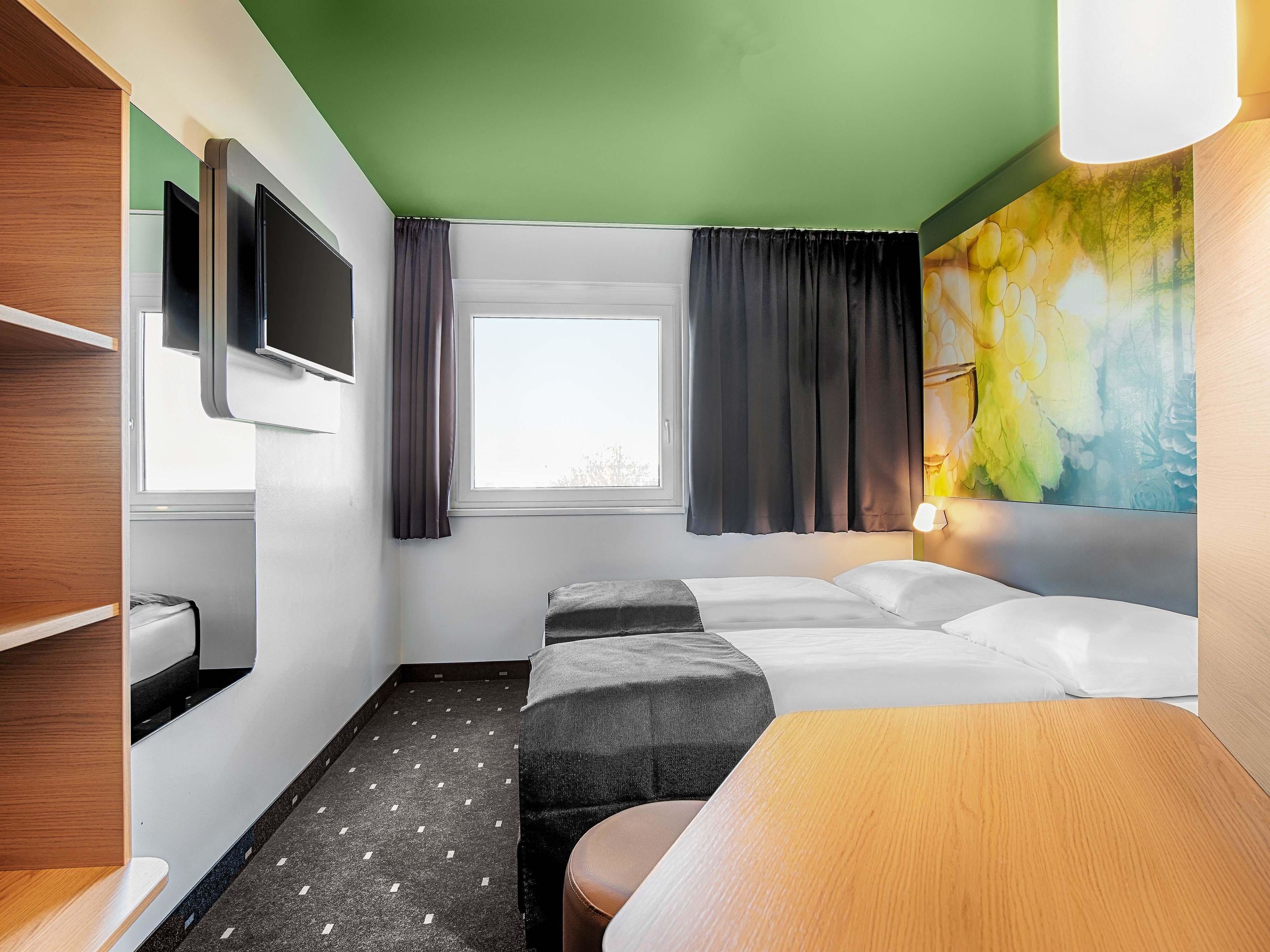 Habitación de hotel con cama, televisor montado en la pared, ventana con cortinas y un cuadro en la pared.