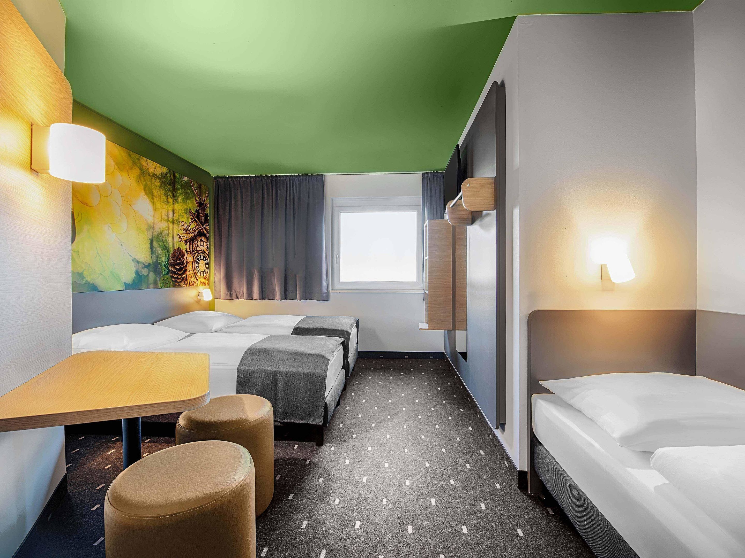 Habitación de hotel con dos camas, una mesita, dos otomanas, una lámpara de pared y una ventana con cortinas.