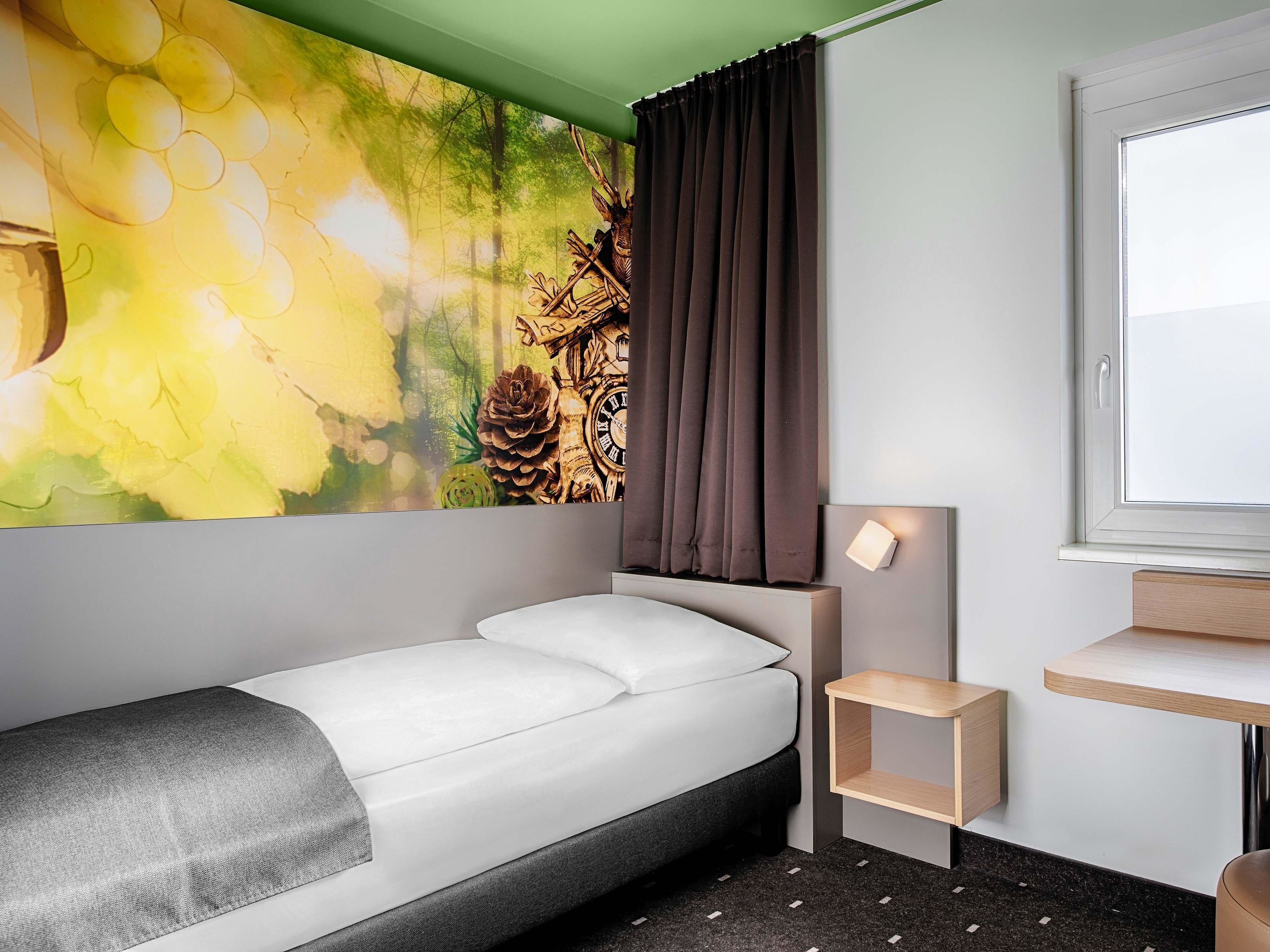 Une chambre d’hôtel moderne, équipée d’un lit, d’une petite table de chevet, d’une lampe fixée au mur et d’une fenêtre donnant sur l’extérieur.