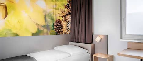 Une chambre d’hôtel moderne, équipée d’un lit, d’une petite table de chevet, d’une lampe fixée au mur et d’une fenêtre donnant sur l’extérieur.