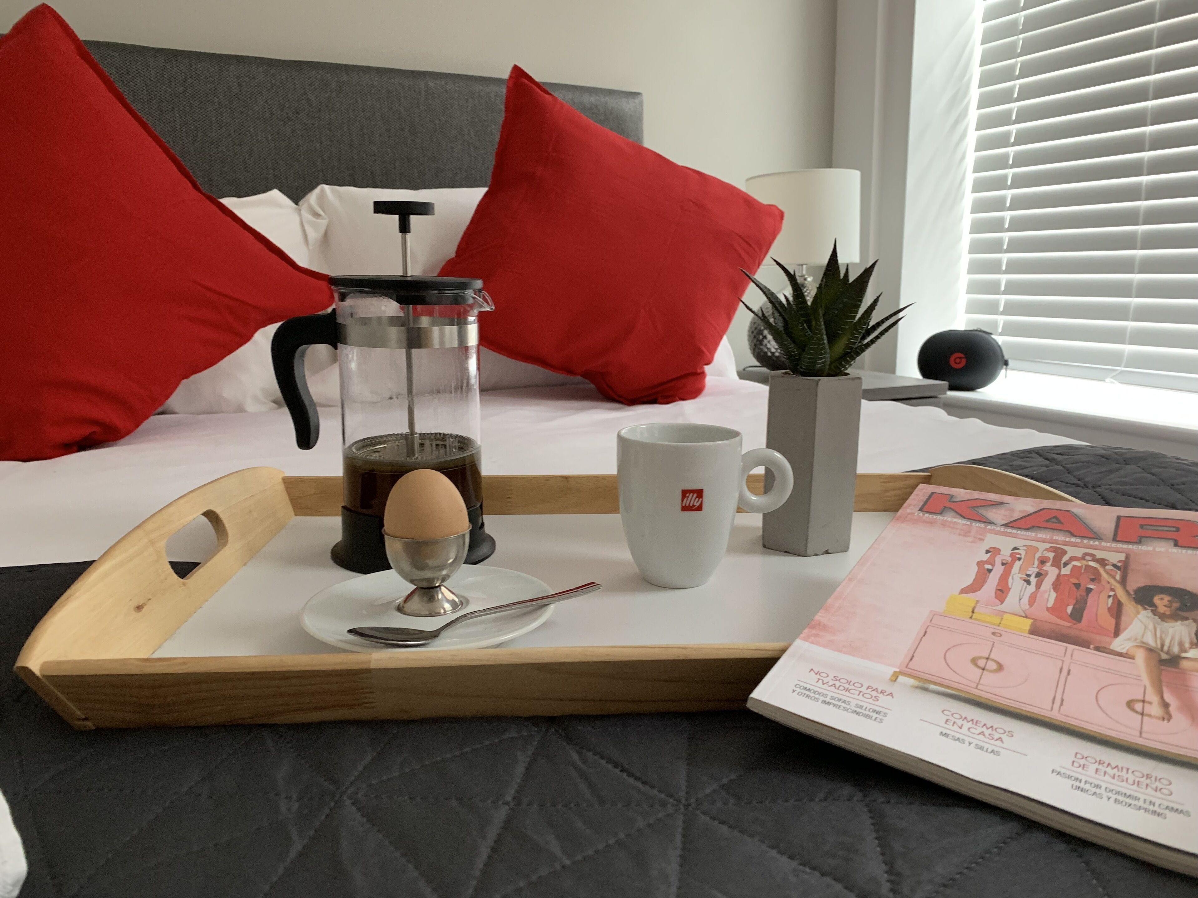 Una cama con almohadas rojas, una cafetera, una taza, una bandeja y una revista sobre una mesita de noche.