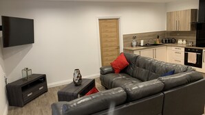 A modern living room with a grey leather sofa, a small black TV stand, and a kitchen area with wooden cabinets and a black range hood. - Impeccable 2-bed Apartment in Birmingham (Birmingham)