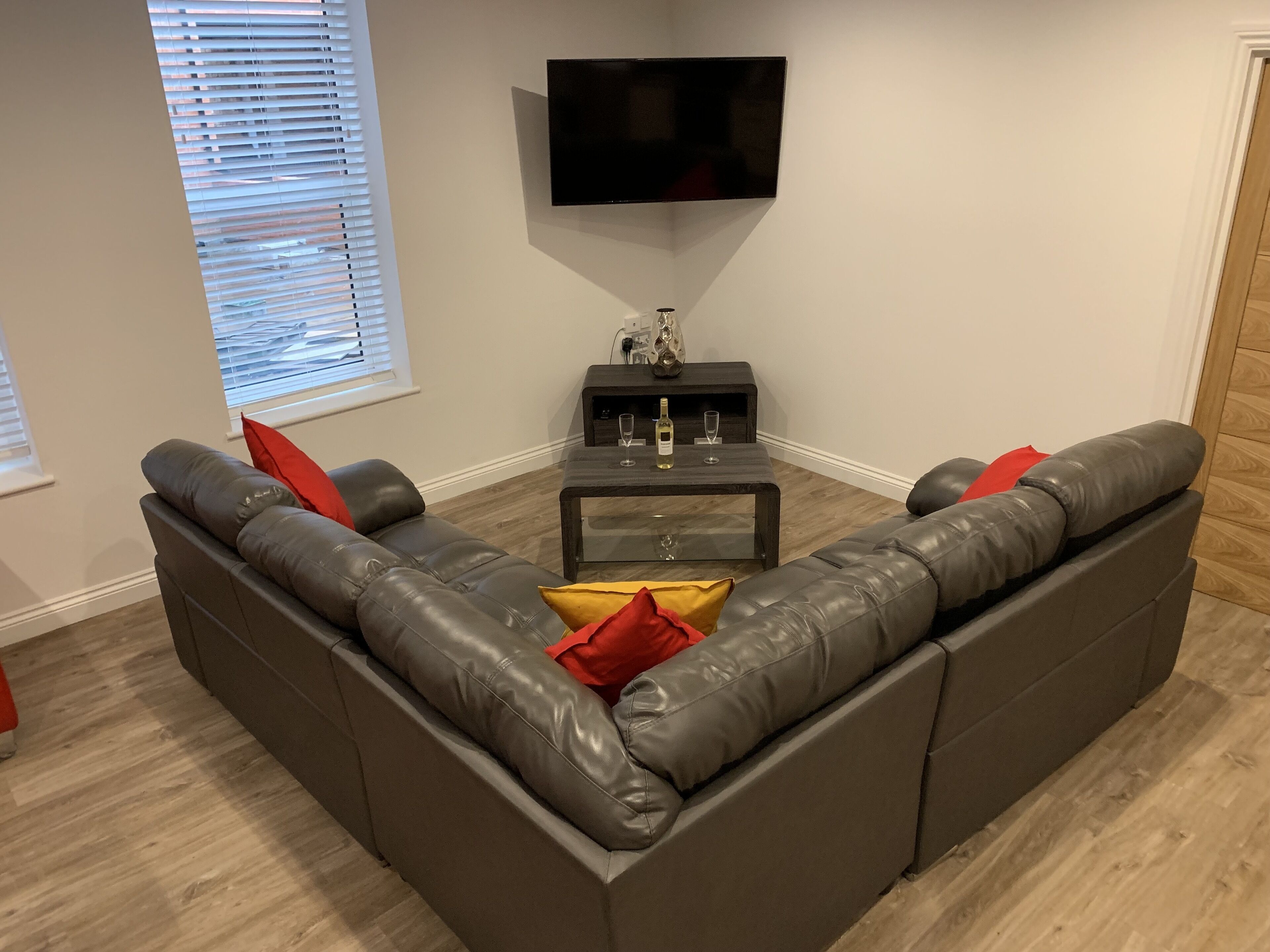 Un salon moderne avec un canapé d’angle en cuir gris, un téléviseur à écran plat fixé au mur et une petite table d’appoint sur laquelle se trouvent une bouteille de vin et des verres.