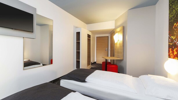A modern hotel room with a bed, a TV mounted on the wall, a mirror, and a red stool.