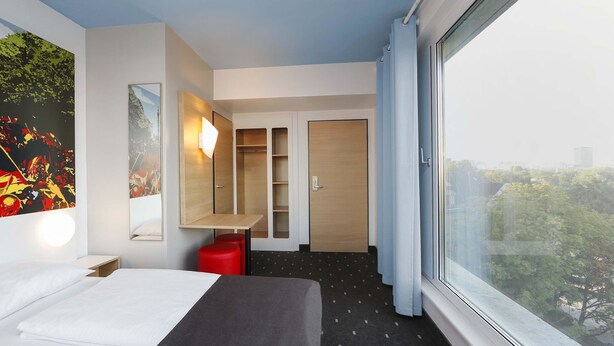 Een moderne hotelkamer met een bed, bureau, kledingkast en een groot raam met uitzicht op de stad.