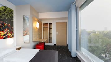 A modern hotel room with a bed, a desk, a wardrobe, and a large window offering a city view.