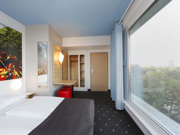 Ein modernes Hotelzimmer mit Bett, Schreibtisch, Kleiderschrank und einem großen Fenster mit Blick auf die Stadt.
