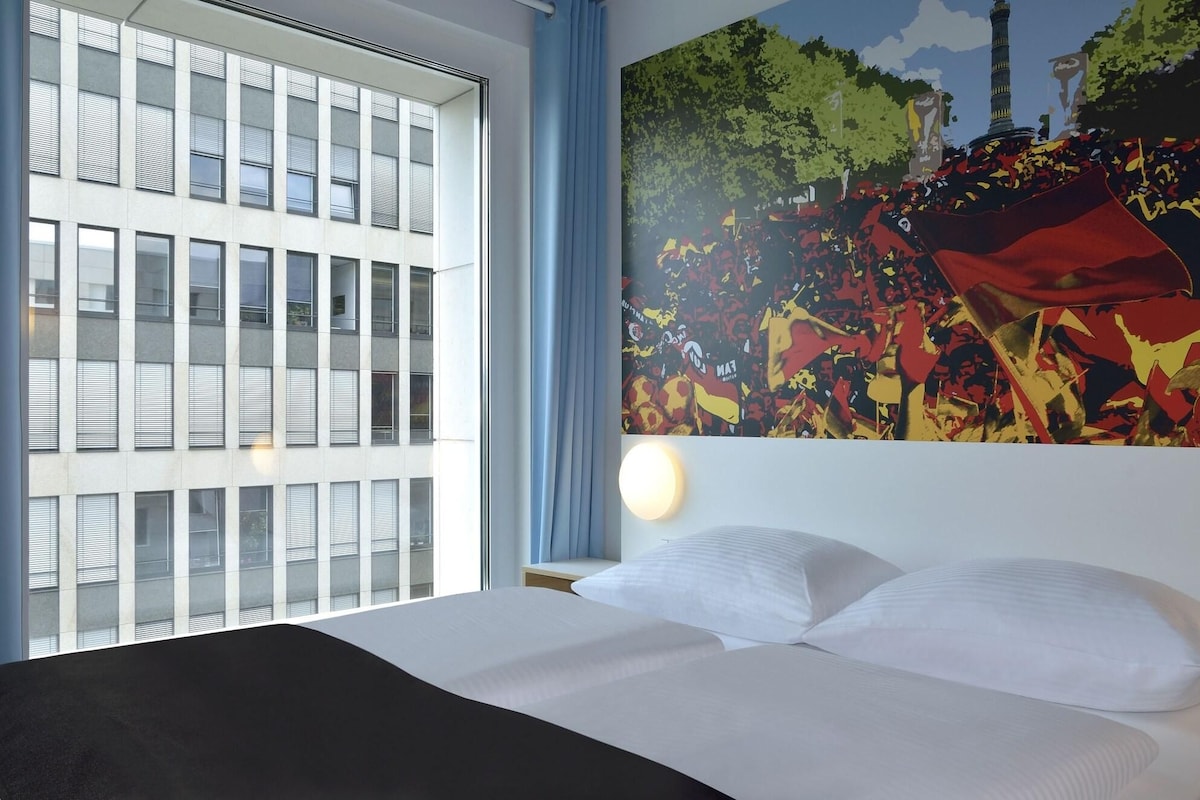 Una habitación de hotel con cama, ventana con persianas y un cuadro abstracto colorido en la pared.