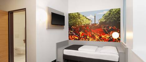 Una habitación de hotel con cama, televisor de pantalla plana montado en la pared y un mural grande que muestra a una multitud junto a un monumento.