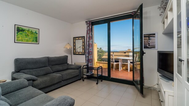 Een moderne woonkamer met een grijze bank, een televisie en een schuifdeur die leidt naar een terras buiten met een tafel en stoelen.